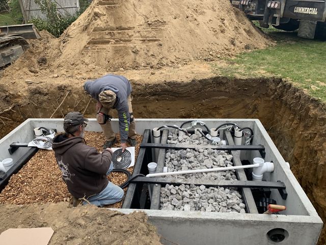 A concrete innovative/alternative septic system sits in a dirt hole in the ground, its lid off. The inside includes two chambers, one side filled with woodchips, the other with limestone.