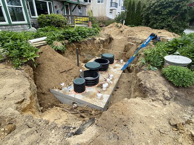 A large concrete septic system sits in a hole in the ground in front of a house, a ladder perched between the tank and the ground.