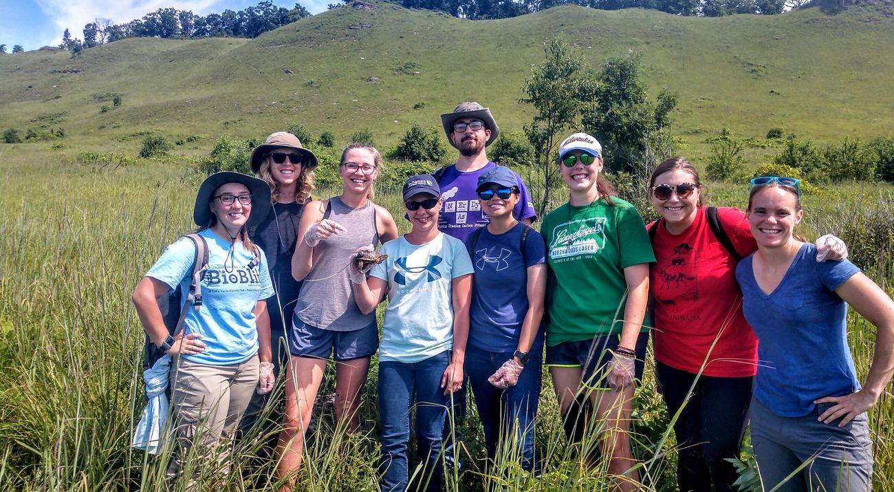 A group of seven interns stand with a background of a tree and wetland stream.