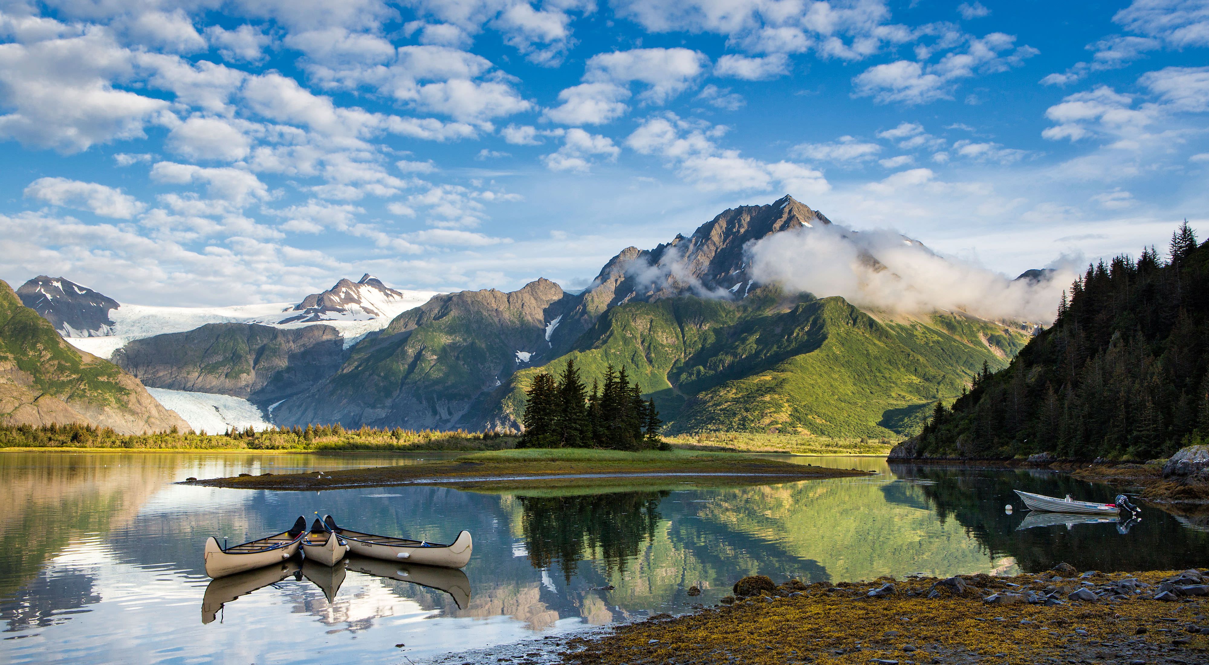Scenic view of a still lake with three canoes floating on it with rugged green and snow-covered mountains in the background under a blue sky dotted with puffy clouds in Alaska.