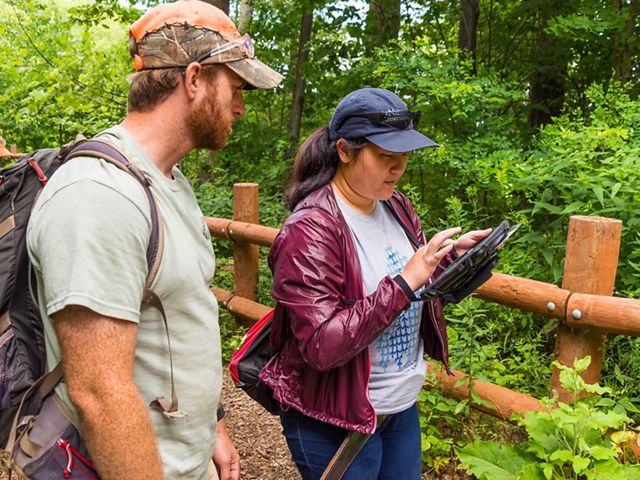 Young woman on trail with blue TNC cap on inputs information into a tablet she holds in her hand while man wearing a backpack observes.