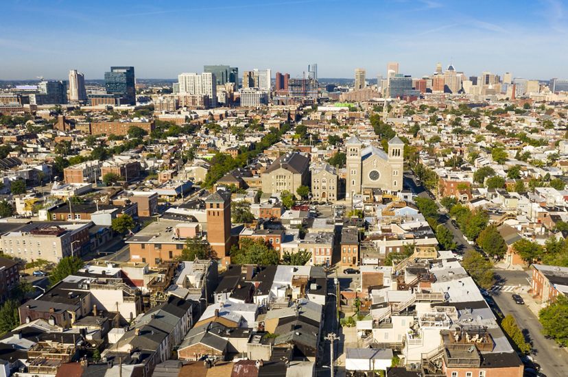 Aerial view of Baltimore looking towards the downtown skyline and Inner Harbor. The foreground is filled with rowhouse lined residential neighborhoods and centered on a large church.