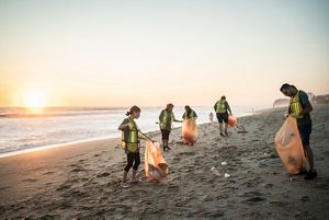 A group of 6 people hold orange trash bags while walking along a beach.