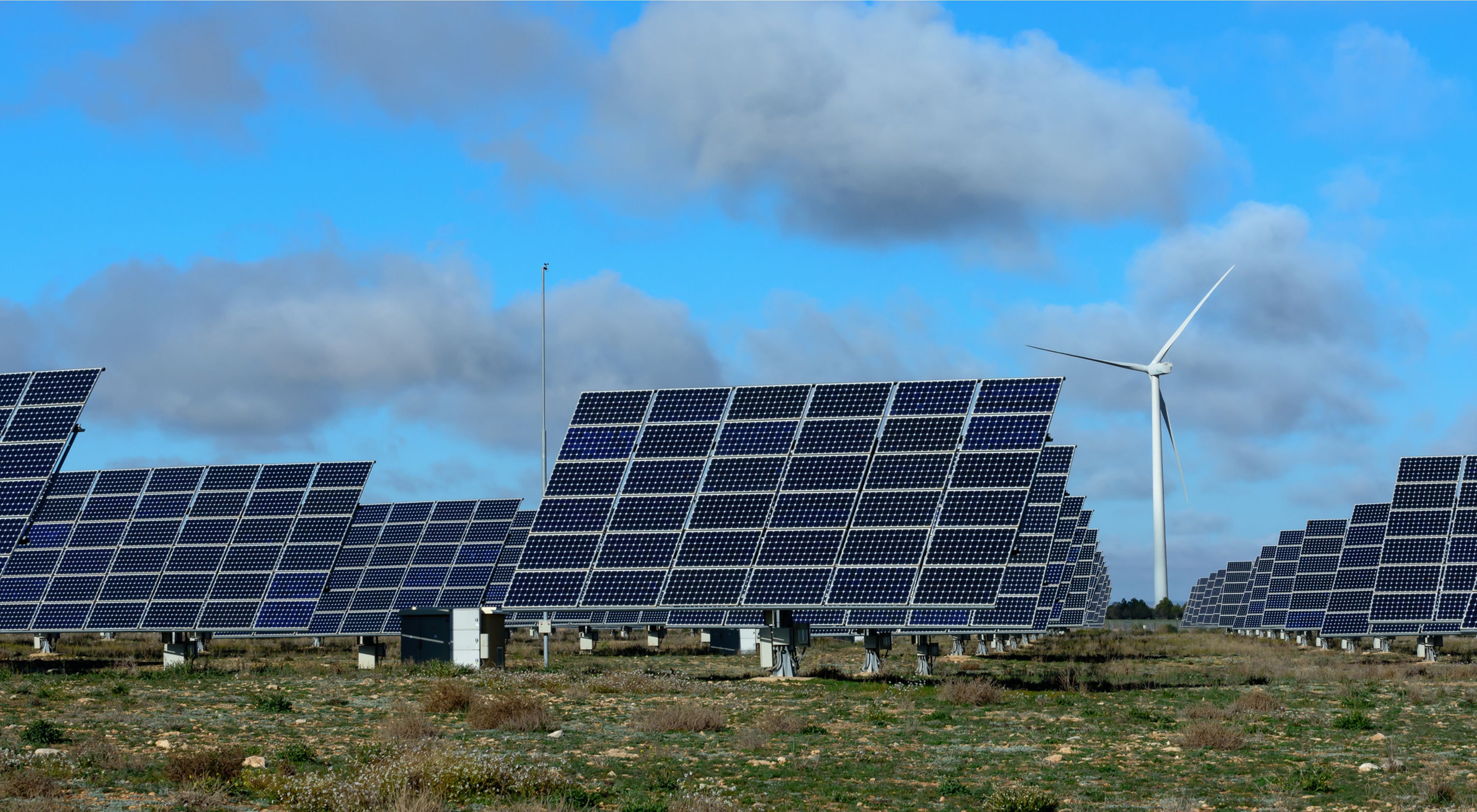 Photo of solar photovoltaic panels and one wind turbine in a field below a bright blue sky.