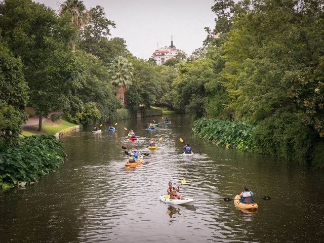 A river, with at least 13 kayakers paddling along, meanders through dense green brush and trees as a skyscraper looms in the distance.