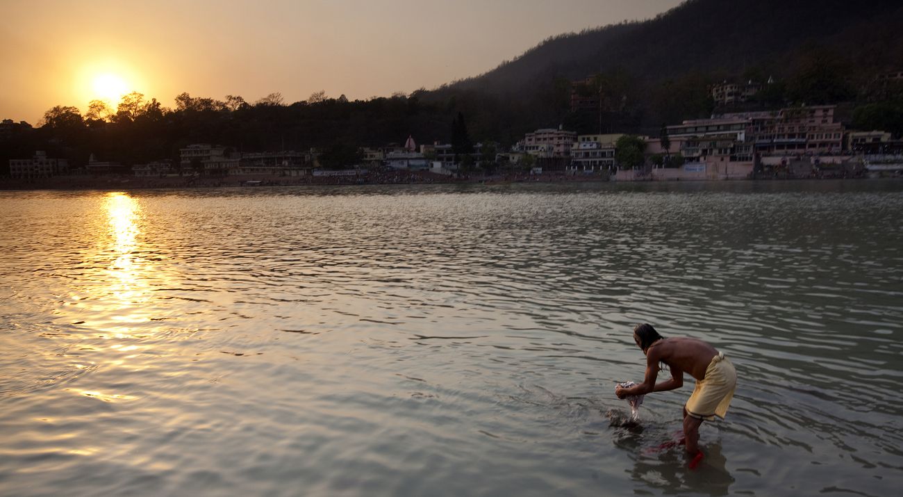 A man bends over and dips his hands in a river as the sun sets on the water.