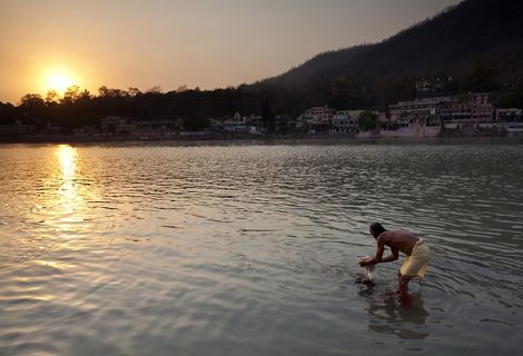 A man bends over and dips his hands in a river as the sun sets on the water.