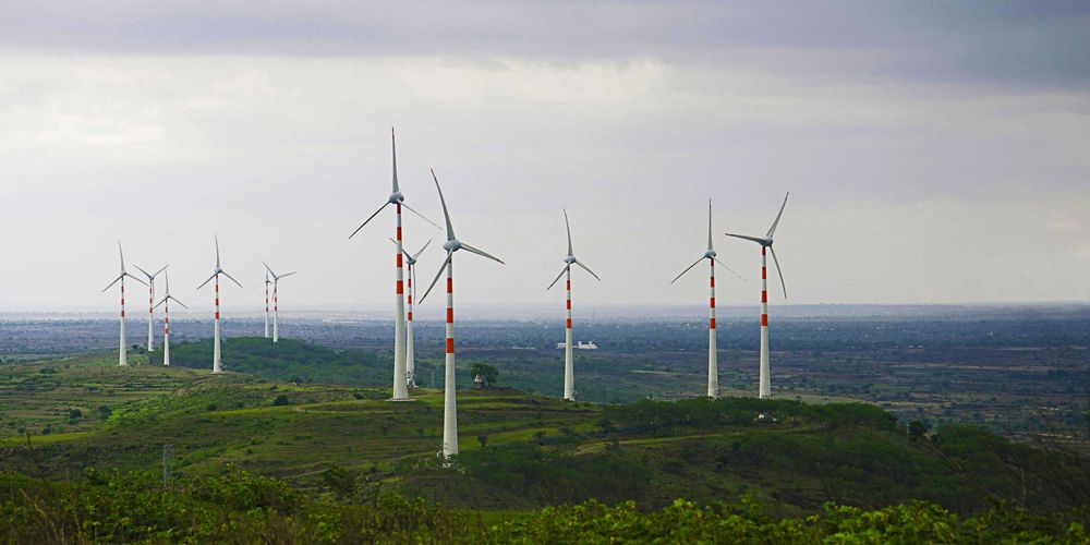 Several white wind turbines with red stripes stand in a vast green landscape in India.