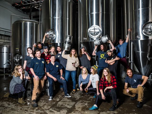 A group of brewery staff members pose near stainless steel barrels.