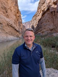 James McGuire stands in front of rocky canyons cut in half by a river lined with green grass.