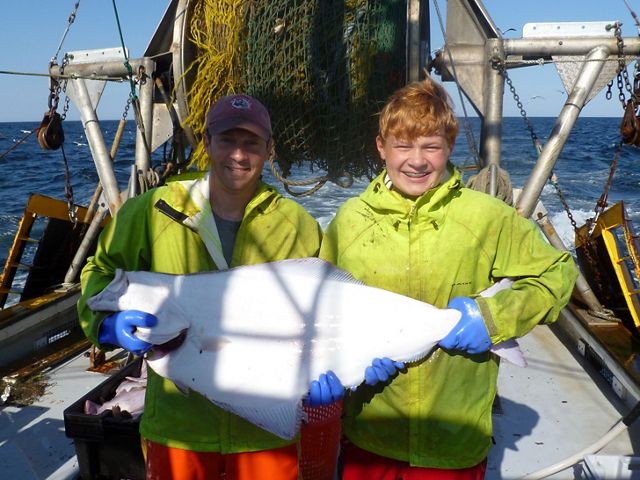 A man and his son standing on the deck of a fishing boat holding a large white halibut.