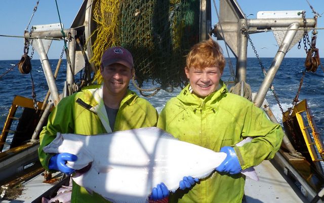 A man and his son standing on the deck of a fishing boat holding a large white halibut.