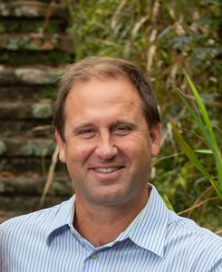 A man with sandy-brown hair and a blue-striped shirt smiles at the camera.
