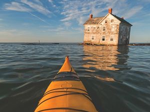 An abandoned farm house sits precariously at the edge of the water as the Chesapeake Bay slowly consumes it. The yellow nose of a kayak is visible in the foreground.