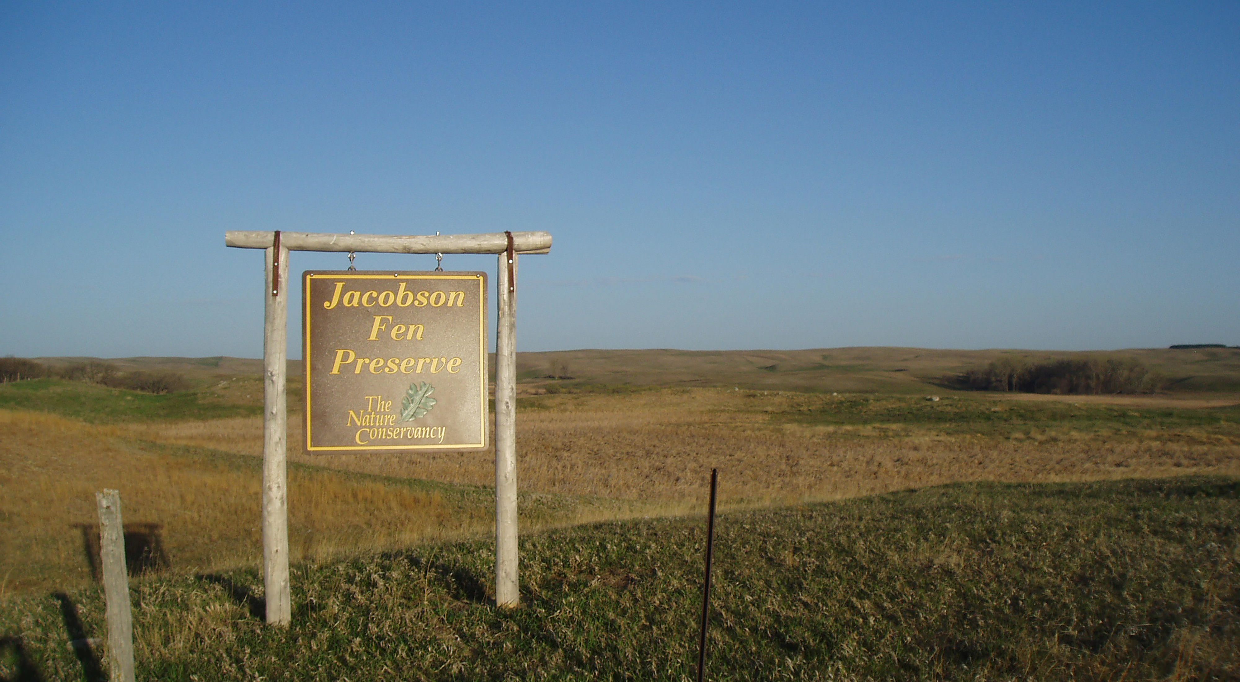 Large sign that reads Jacobson Fen Preserve with rolling prairie landscape in the background.