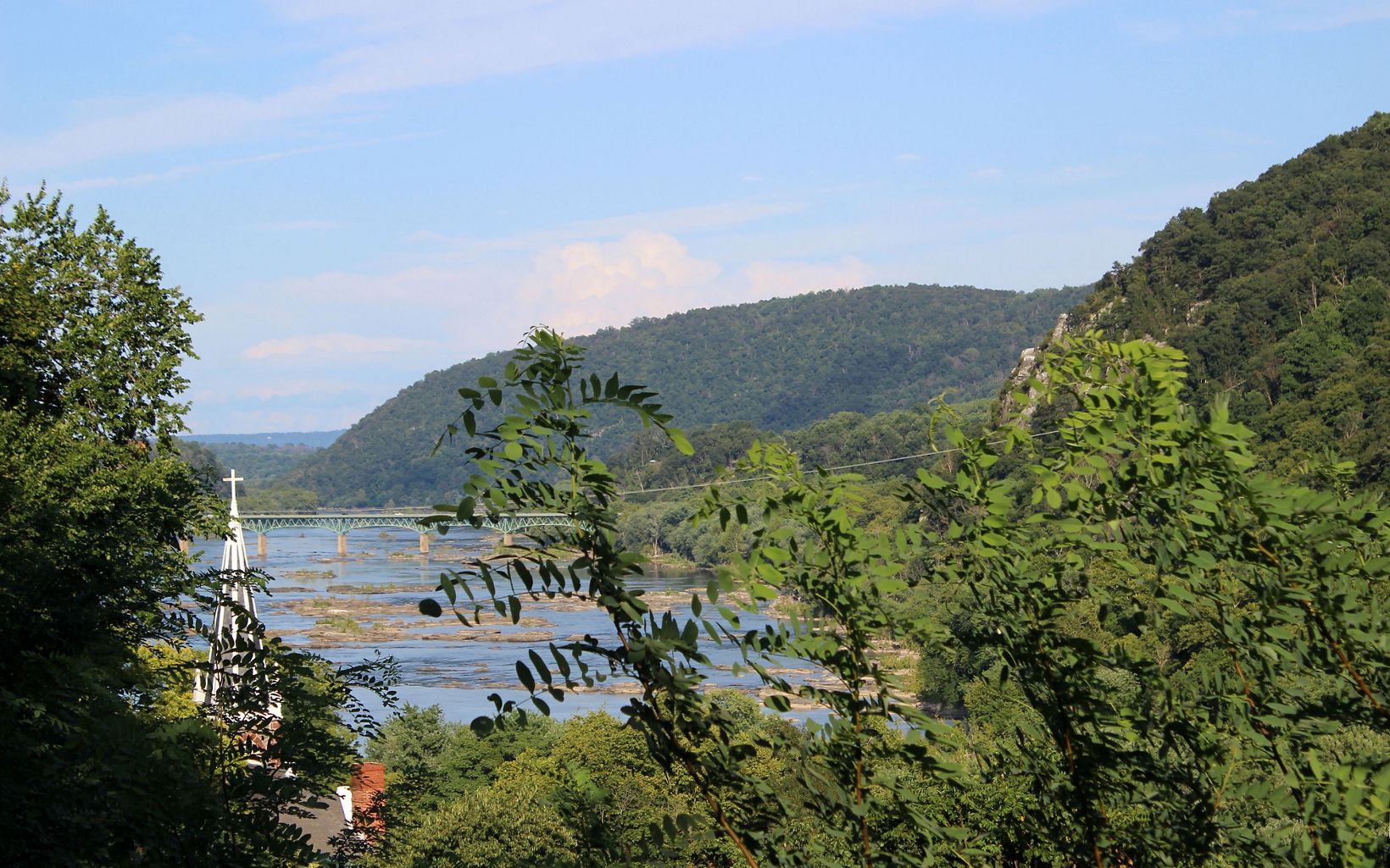 View through tree branches of the Potomac River as it winds between green mountains and the town of Harpers Ferry.