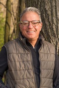 A photo of a glasses-wearing man with short grey hair standing outside next to a tree. 