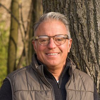 A photo of a glasses-wearing man with short grey hair standing outside next to a tree.