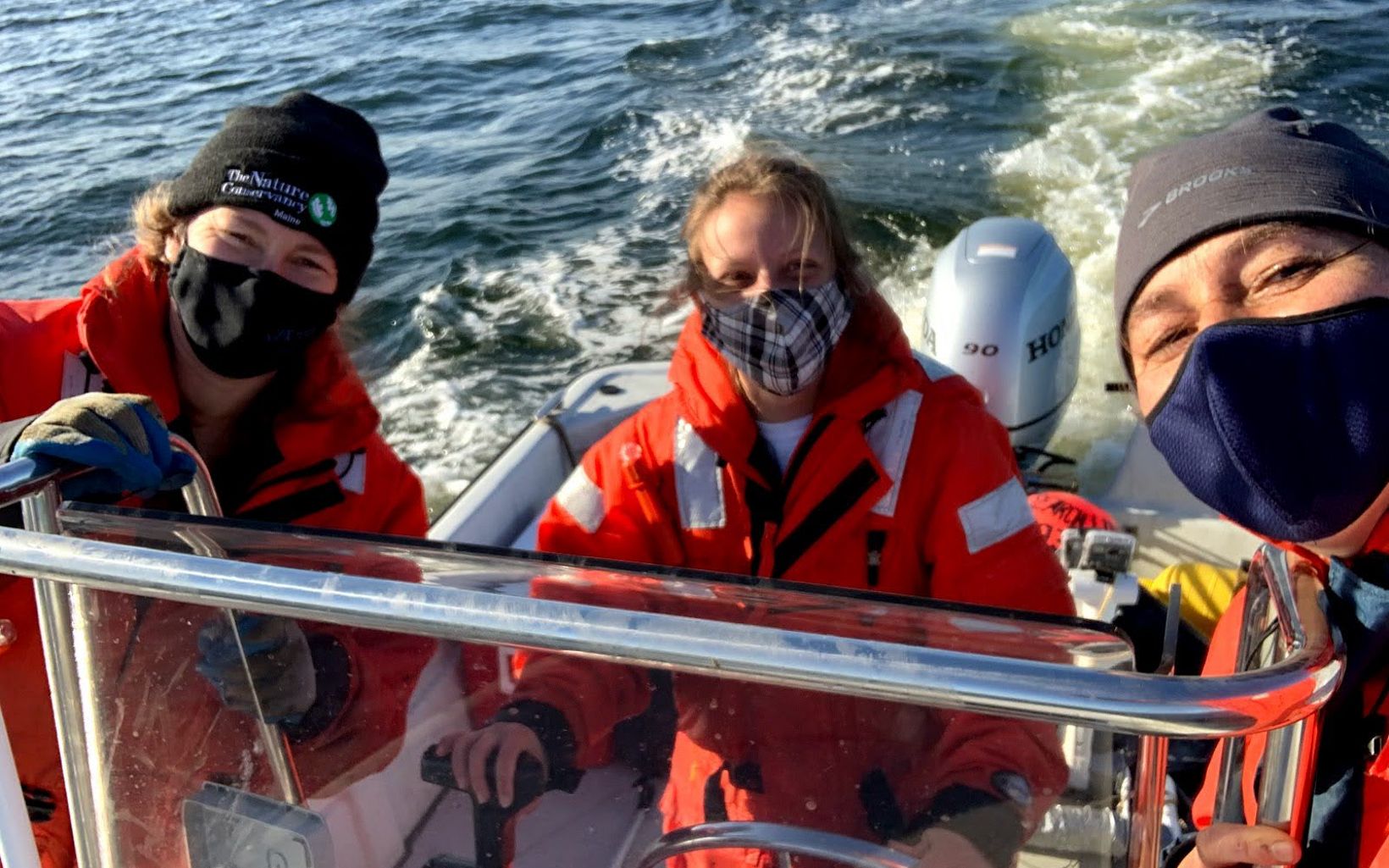 Three women in dry suites stand in a moving motor boat.