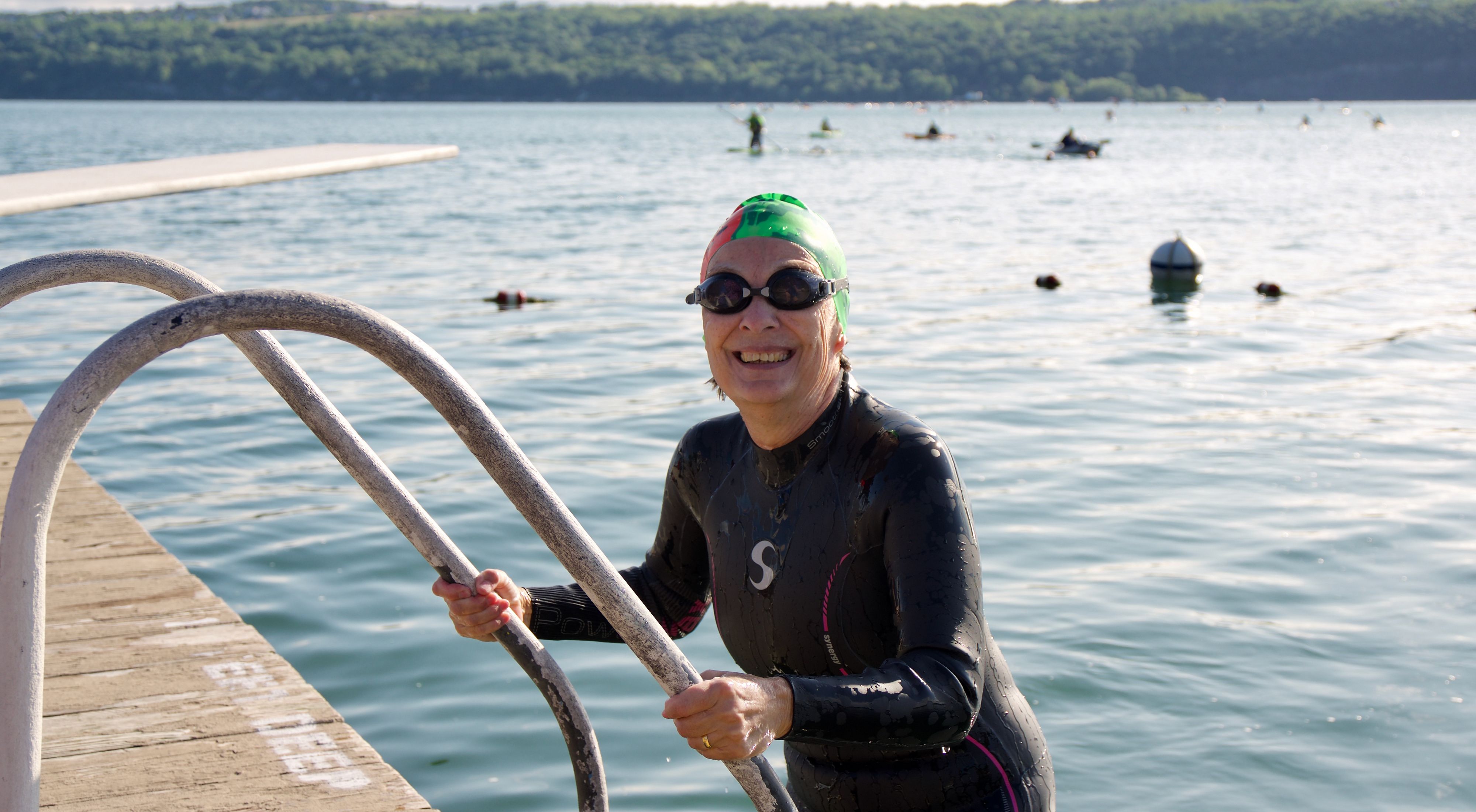 A swimmer in a long sleeved black wetsuit and green swim cap with goggles on emerging from a lake on a ladder and dock at the lefthand side of the frame.
