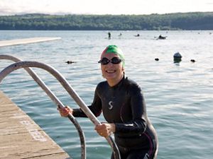  A swimmer in a long sleeved black wetsuit and green swim cap with goggles on emerging from a lake on a ladder and dock at the lefthand side of the frame.