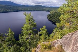 A view from atop a gray boulder on the righthand side of the image looking out onto a blue lake with green trees lining the shore.