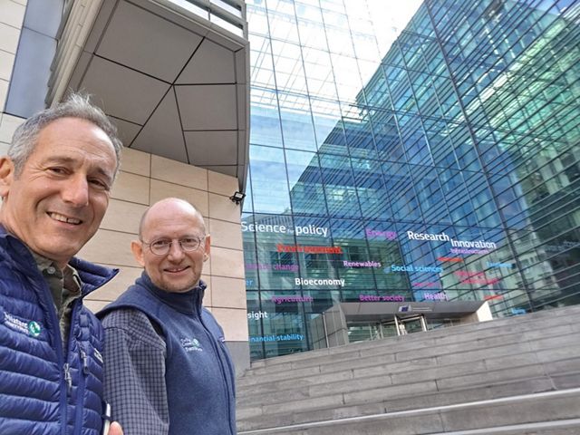 Josh Royte and Andras Krolopp in front of a glass-walled building with words like "science", "policy", "innovation" on it.