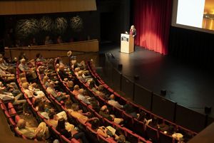 A woman standing in a theater at a podium talking to a room full of people.