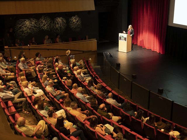 Person standing on a stage speaking to a room of people.
