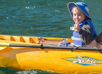 A young child sits in a yellow kayak on a body of water and smiles and gives a thumbs-up to the camera.