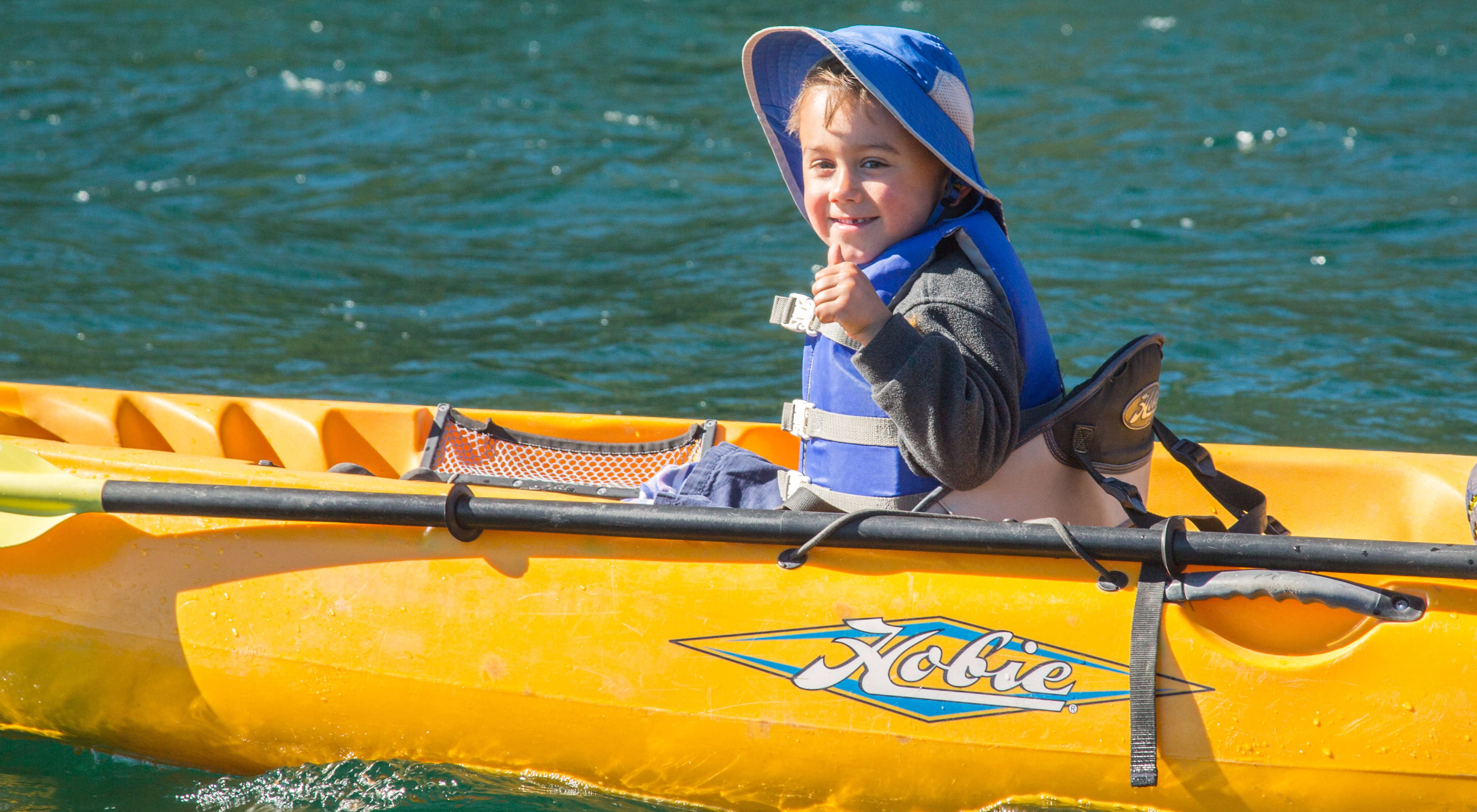 A young child wearing a hat and a life vest sits in a yellow kayak in blue water.