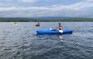 Three kids paddle kayaks of different colors in a lake.