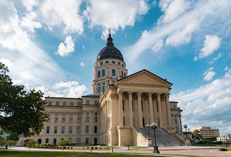 The Kansas State Capitol, a domed building with six stone columns at its entrance, below a partly cloudy blue sky.