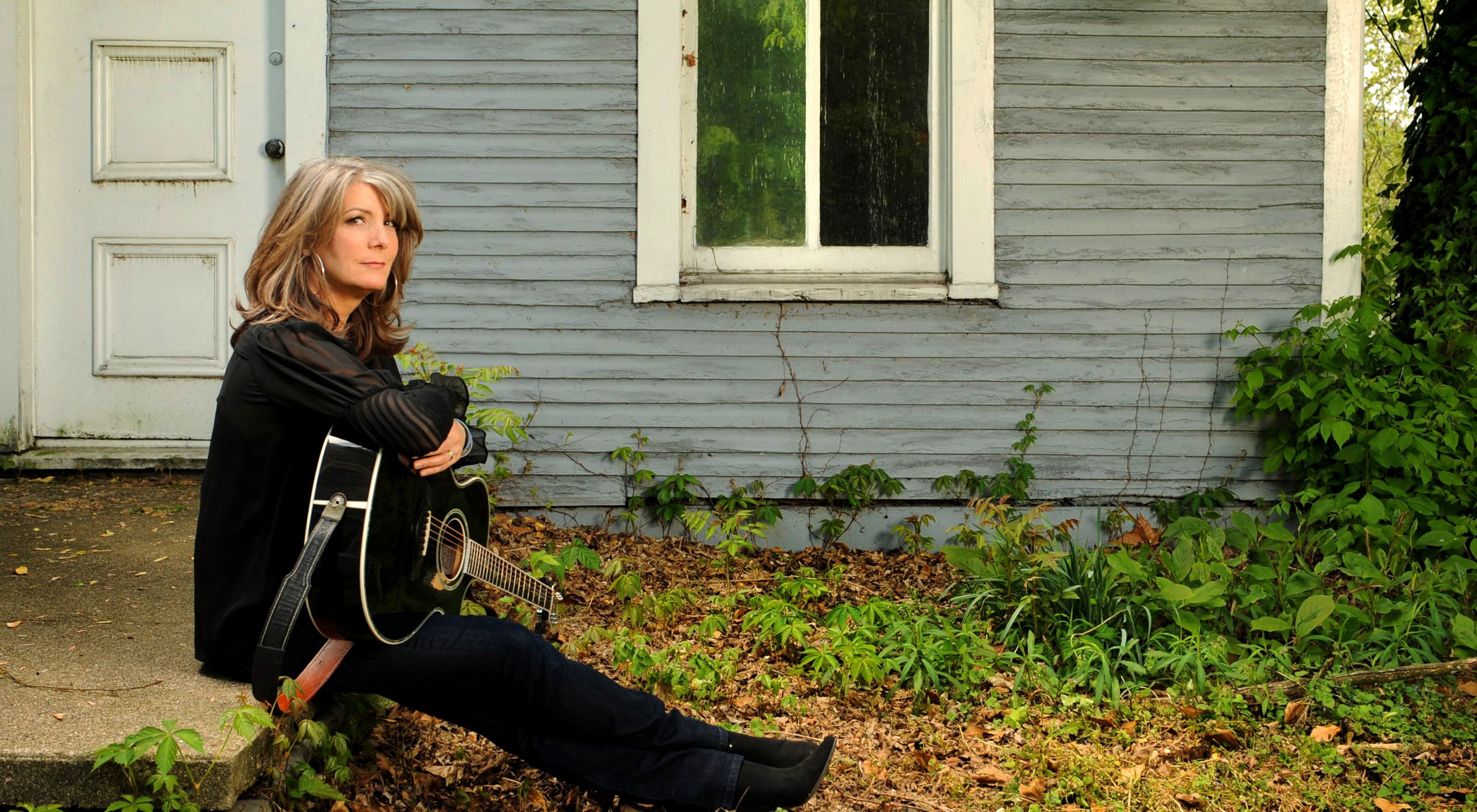 Kathy Mattea holding a guitar in her lap and sitting on a concrete walkway in front of an old house.