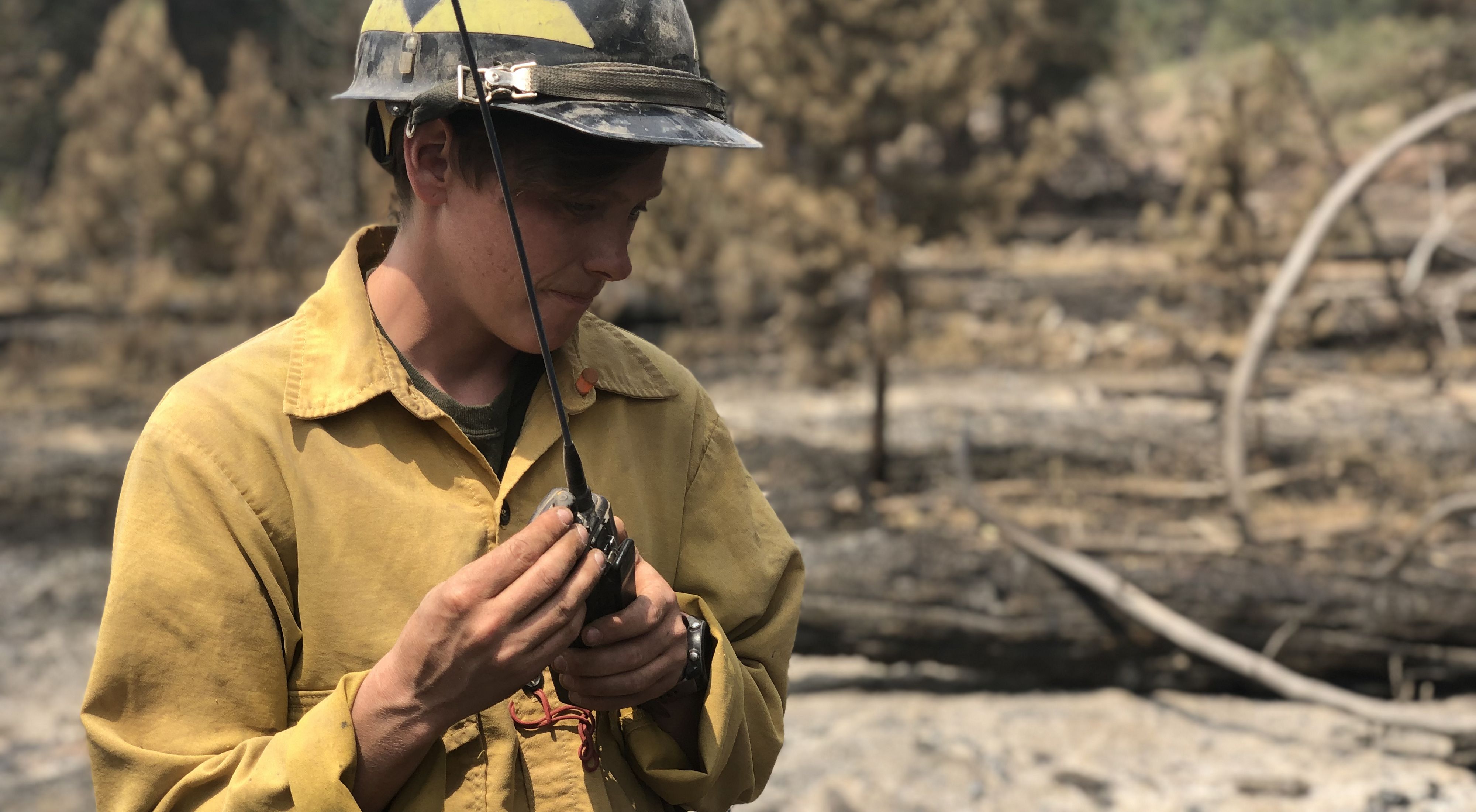 TNC Oregon Fire Program Director Katie Sauerbrey stands in a forest and listens to a walkie talkie; she is wearing fire-protective clothing and a hard hat.