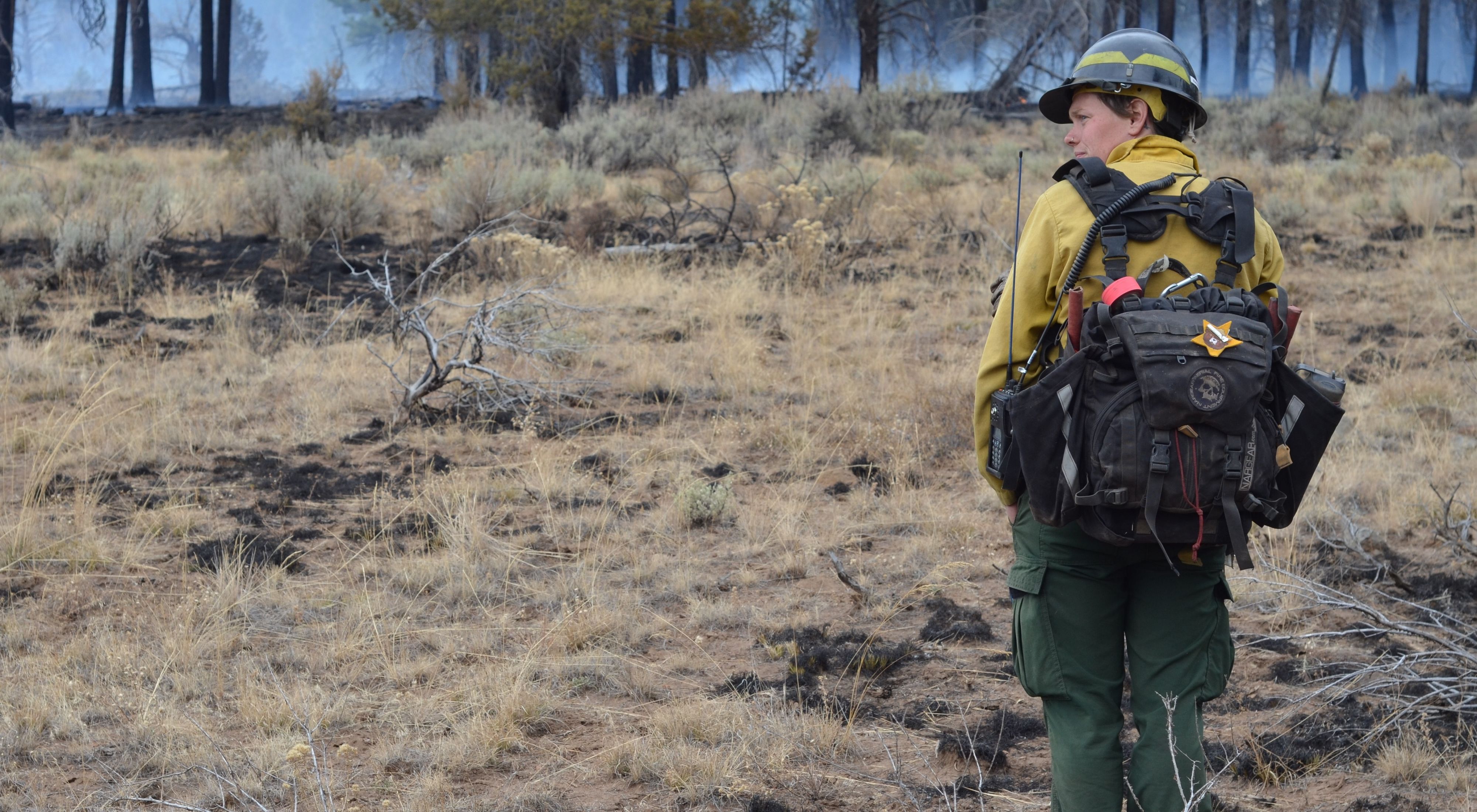 TNC Oregon Burn Boss Katie Sauerbrey stands in fire practitioner gear with their back turned and a forest in front of her.