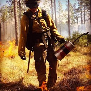 Katie Sauerbrey in fire gear holding a torch with trees and flames behind them.