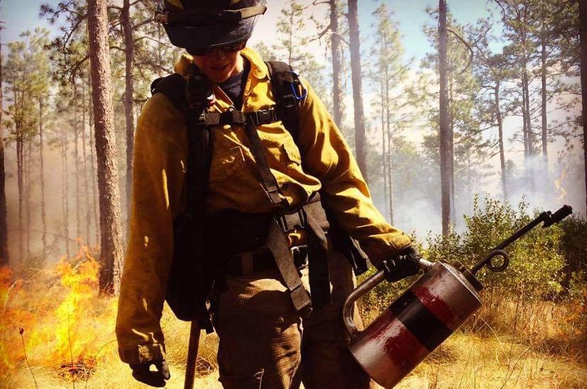 Katie Sauerbrey in fire gear holding a torch with trees and flames behind them.