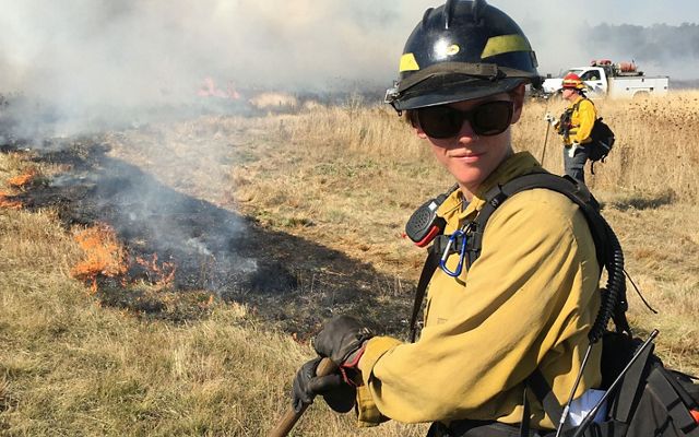 Katie Sauerbrey in fire gear standing in front of a line of burned, blackened grass.