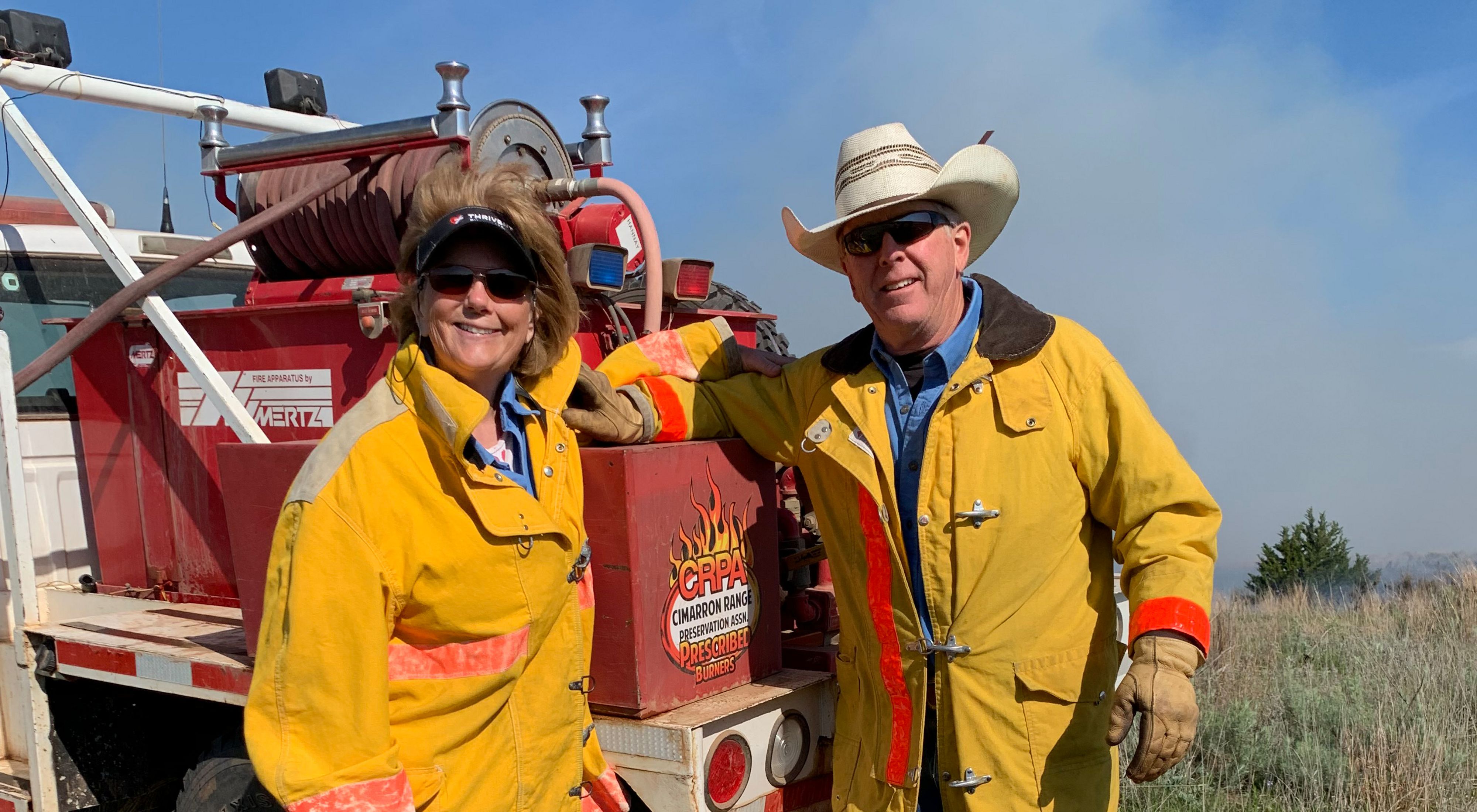 Man and women dressed in protective fire gear stand in front of a utility truck during a prescribed burn on a ranch.