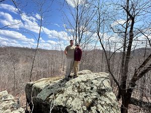 Two people stand on a rock outcropping which overlooks land in a valley below. 