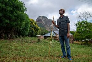 A farmer stands holding a wooden tool with building and a mountain in the background.
