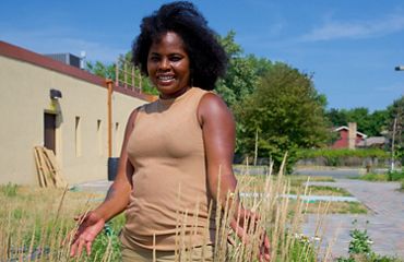 Kenya sitting cross-legged in a grassy area with a row of rocks in the background with her eyes closed and a gentle smile.