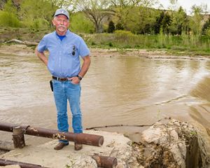A farmer stands in front of a muddy canal as it spills over a small waterfall.