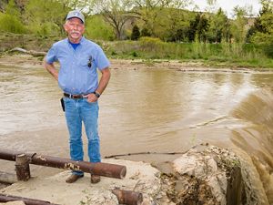  A farmer stands in front of a muddy canal as it spills over a small waterfall. 