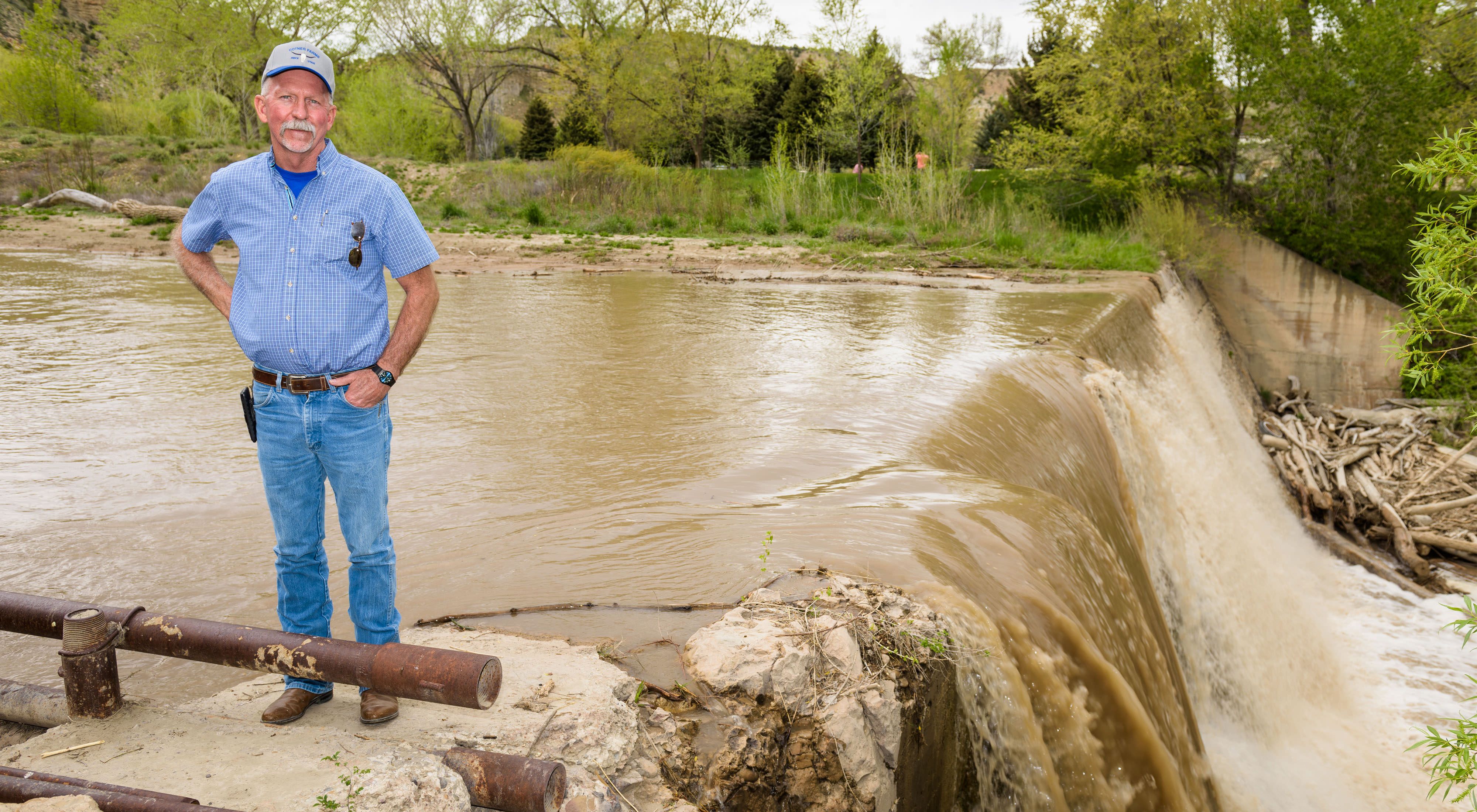  A farmer stands in front of a muddy canal as it spills over a small waterfall. 