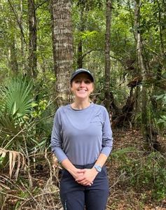 A woman with a baseball cap stands in front of a verdant forest.