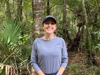 A woman with a baseball cap stands in front of a verdant forest.