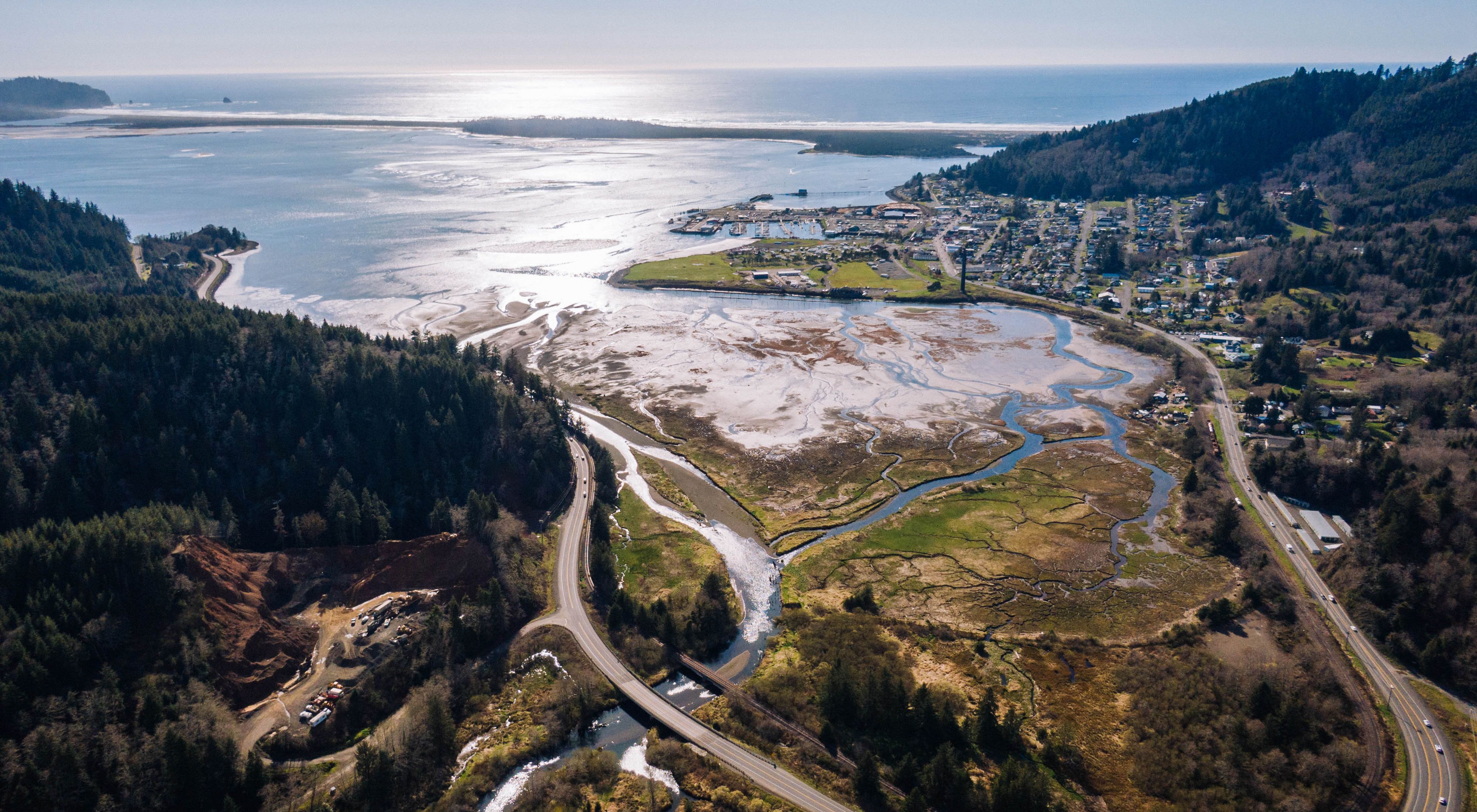 Aerial view of an estuary with meandering and interconnecting streams flowing into a coastal bay, between a town and forests, all along a coastal highway.