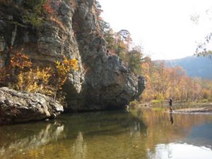 A man is fishing in a calm stream near a large rock bluff. 